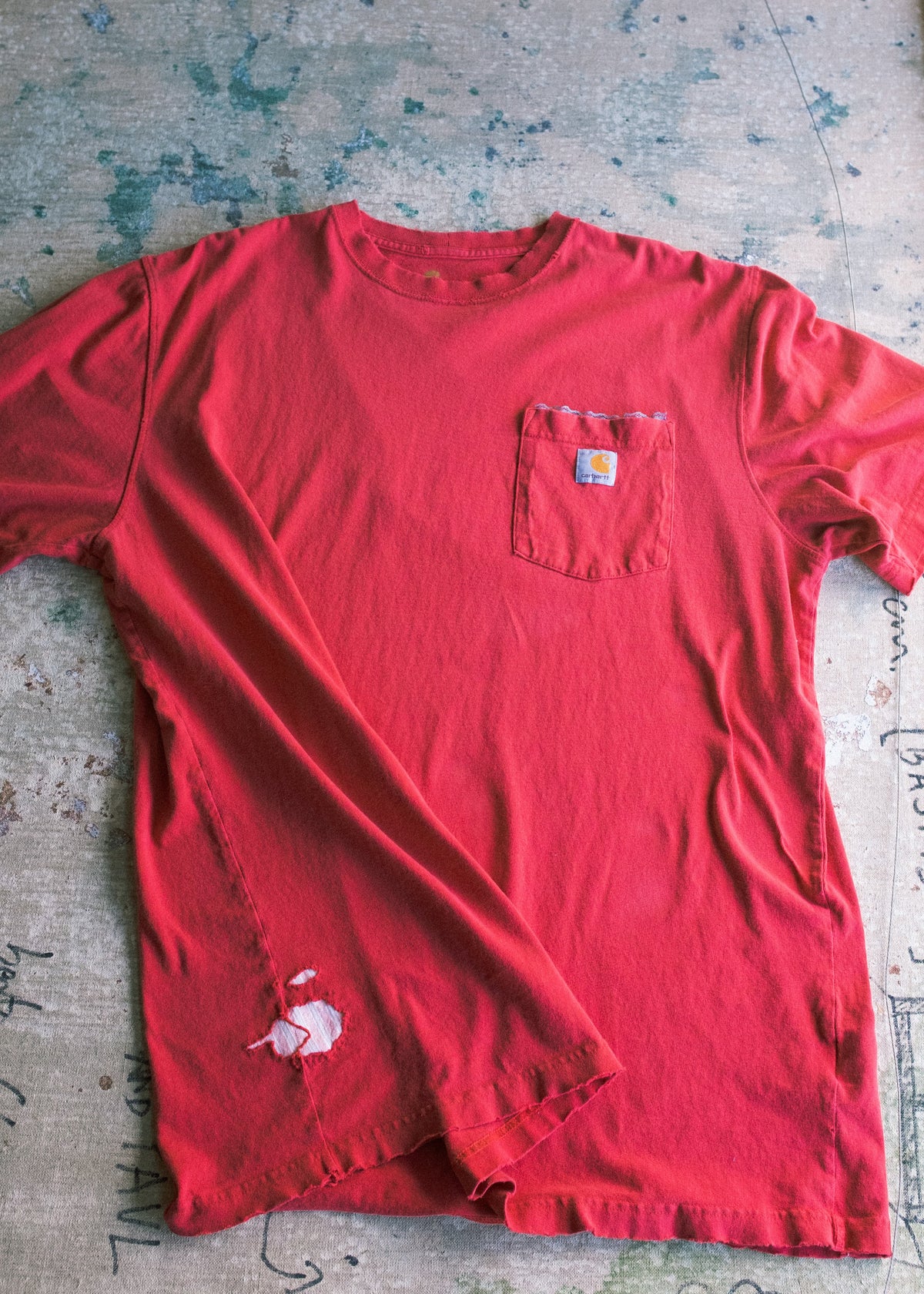 A red short-sleeve pocket t-shirt with a small Carhartt logo patch on the chest pocket, laid flat on a textured concrete surface.